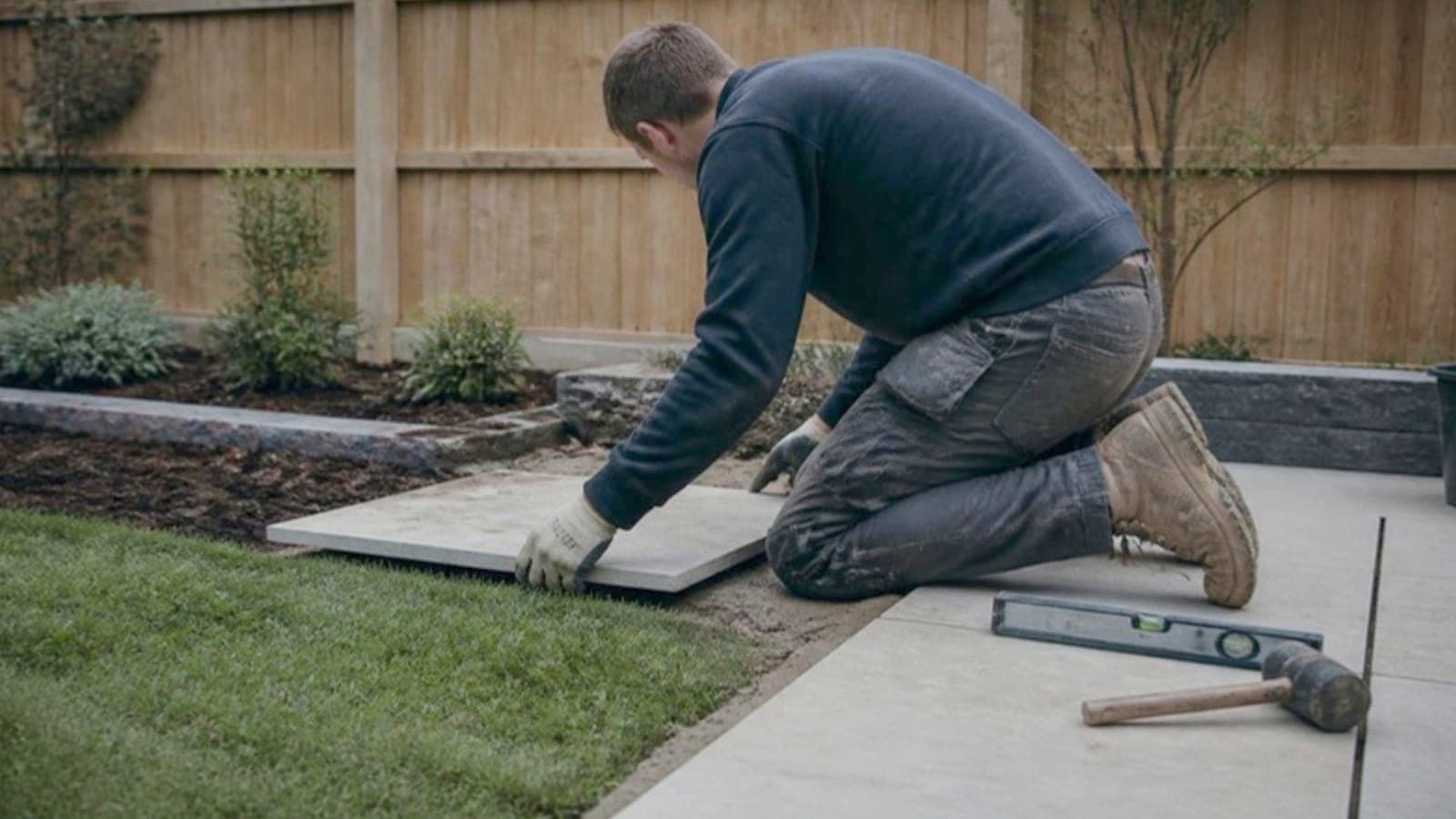 A landscaper laying a porcelain paving slab on a new garden terrace beside fresh turf